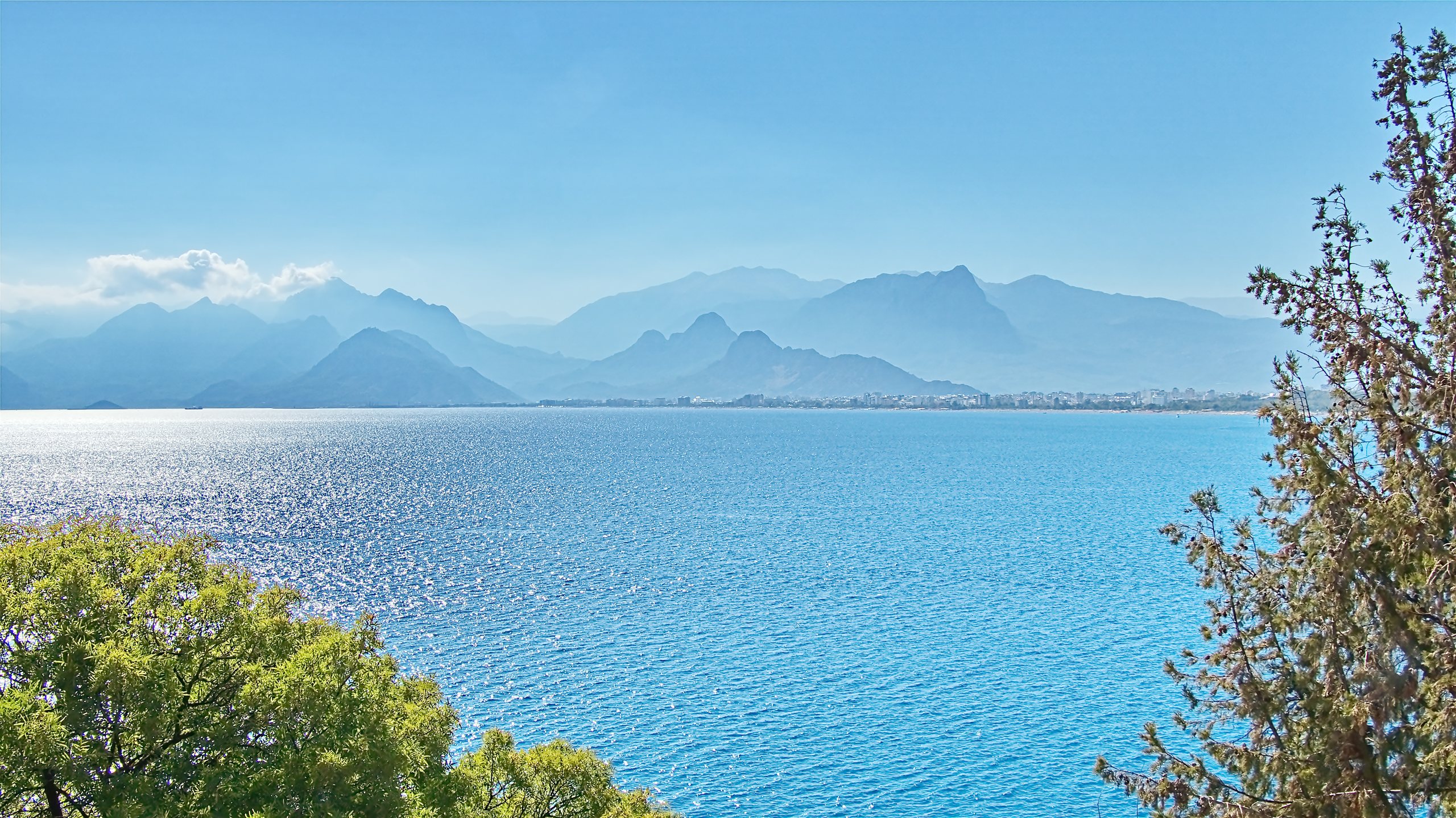 The Gulf of Antalya from the hills: turquoise water, glittering sunlight, the city on the far shore and the hazy Taurus mountains across the bay.