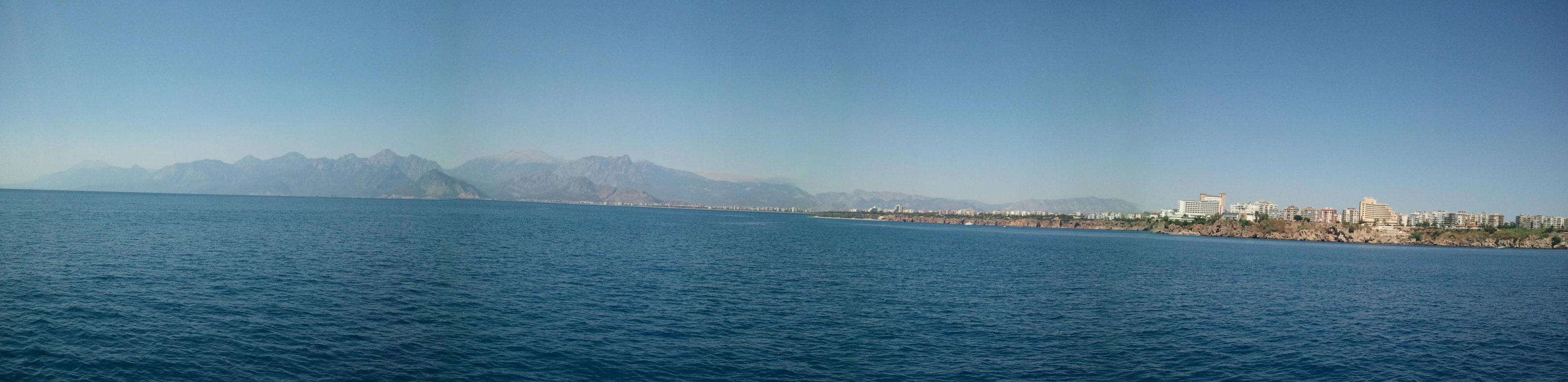 Panorama of Antalya seen from the sea: the old town and its minarets on the cliff edge above the water, masts in the bay, mountains fading into haze.