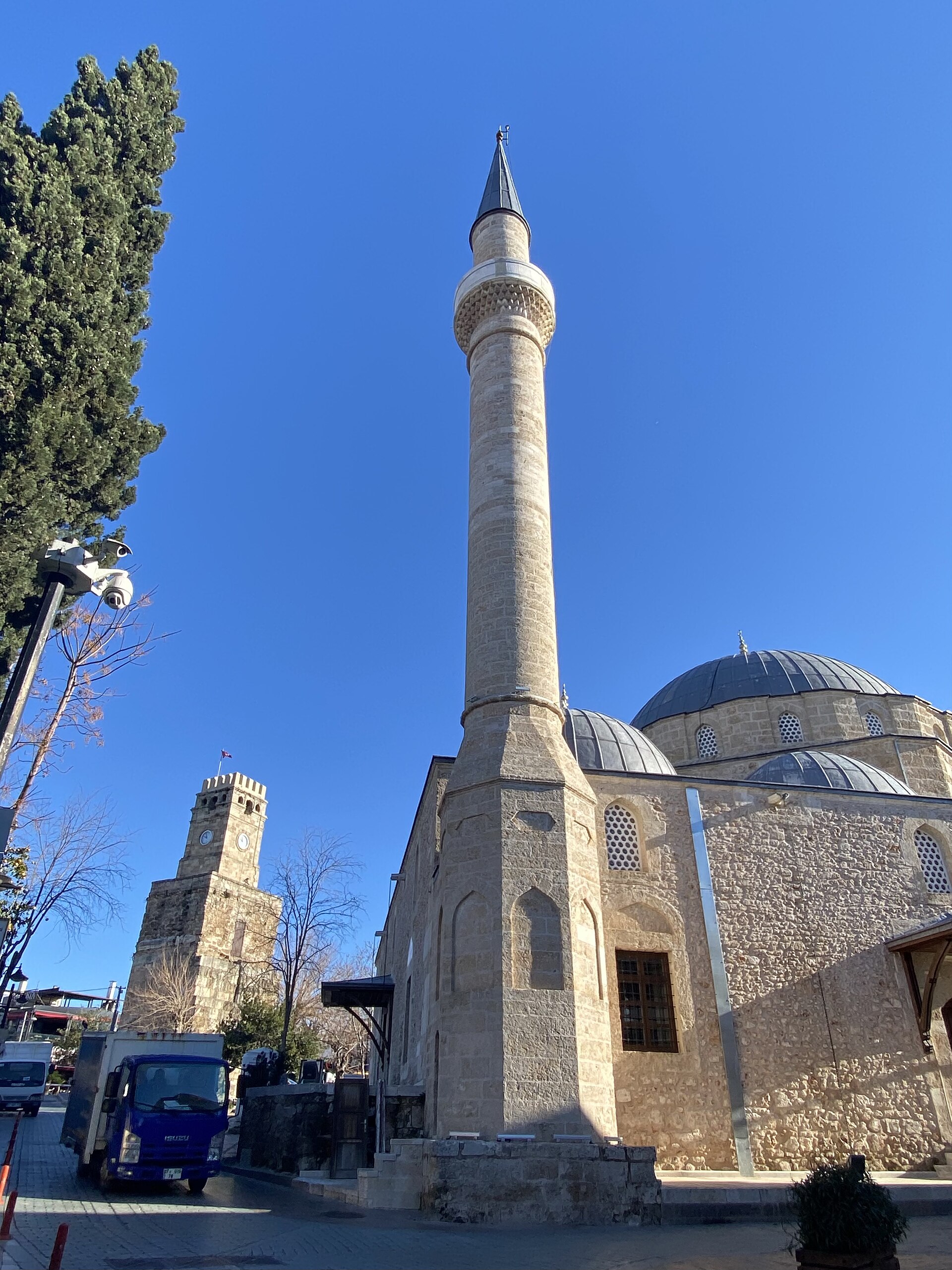 The Antalya Clock Tower, a square stone tower with clocks on each face and a small dome, beside the domes and slender pencil minaret of the Tekeli Mehmet Paşa Mosque against a deep blue sky.