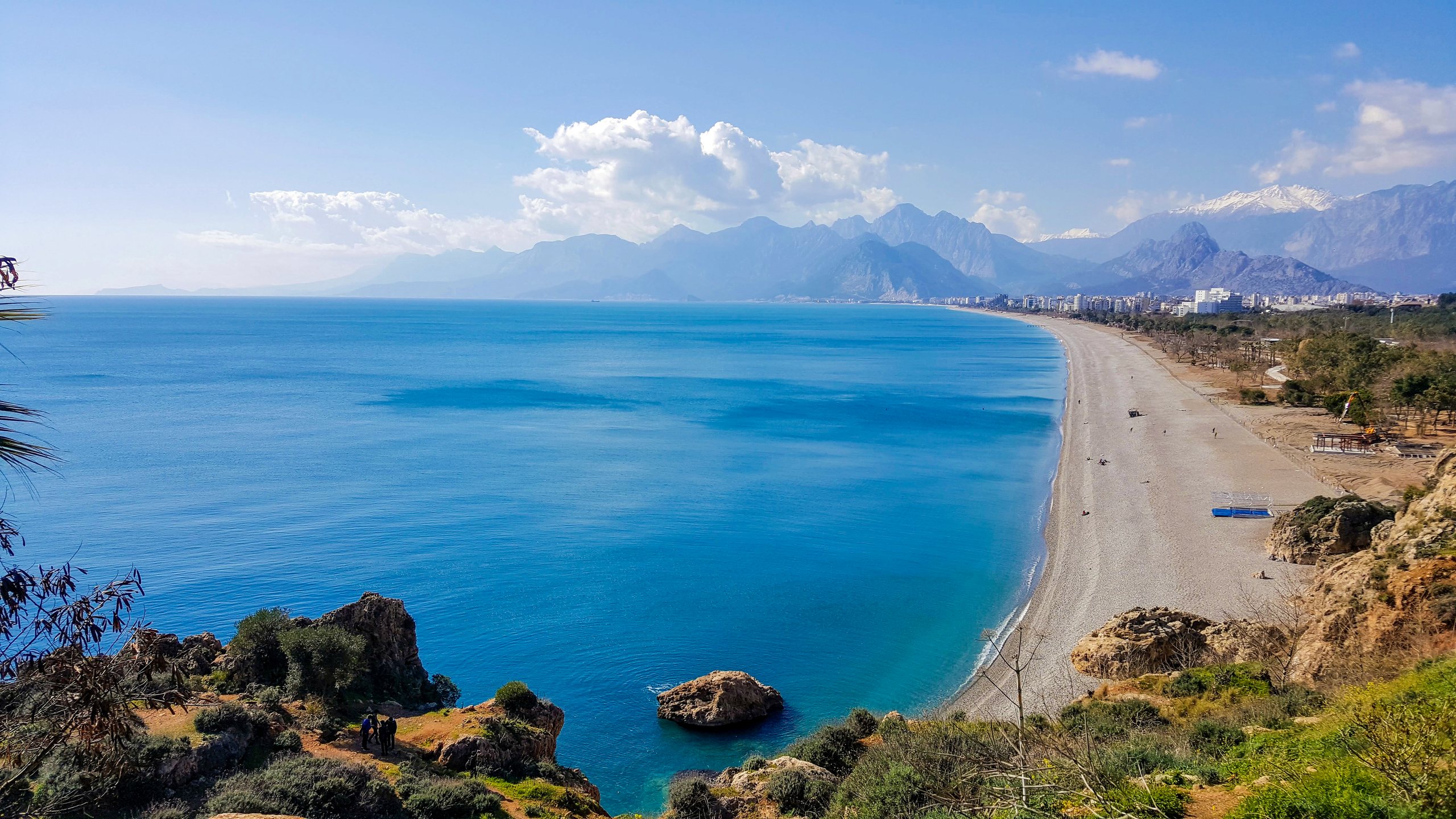 View from the falez cliffs across the rooftops of Antalya toward Konyaaltı Beach and the bay, the sea bright under a clear sky.
