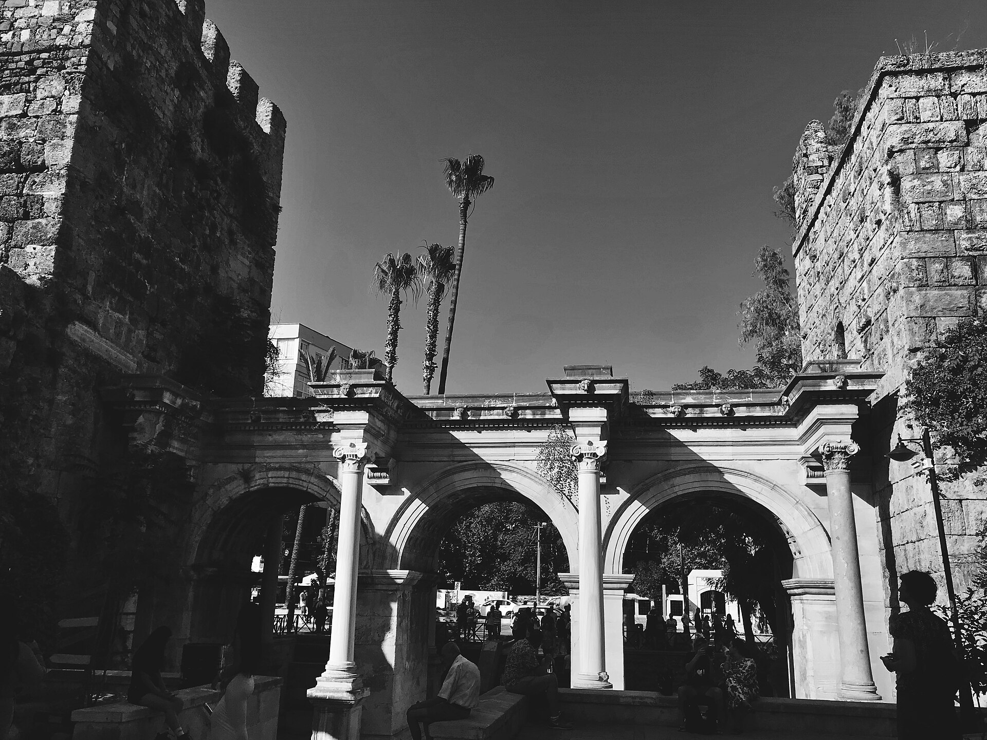 Hadrian's Gate in Antalya, seen in black and white from within the archway: three marble arches with Corinthian columns, weathered stone towers on either side, palm trees and sky above, the old town visible through the arches.