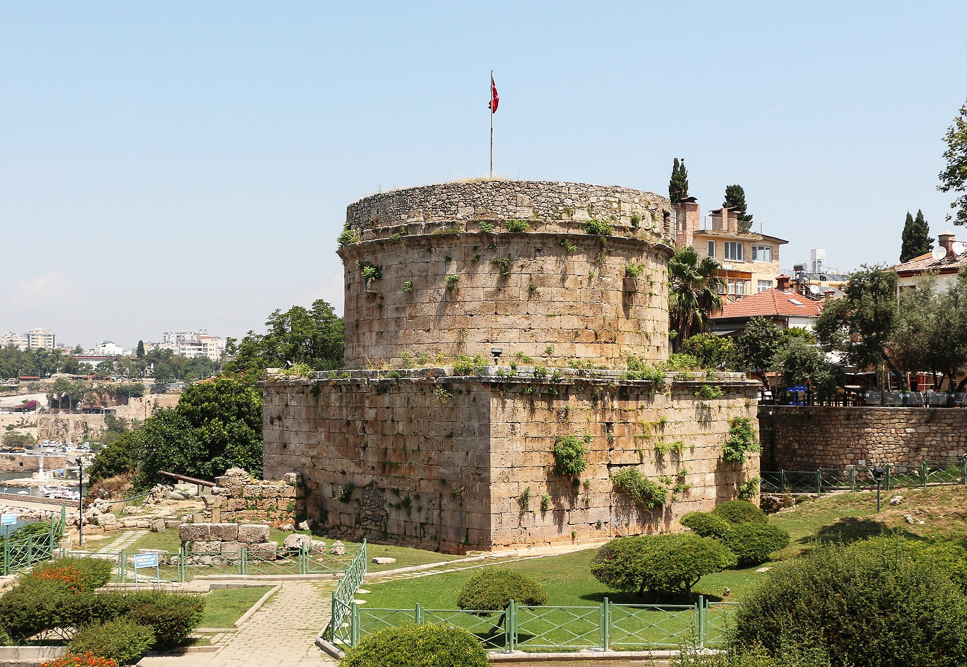The Hıdırlık Tower, a round Roman-era stone tower on a square base at the edge of Kaleiçi's old sea walls, in a small park with the red roofs of the old town behind and the modern city hazy beyond.