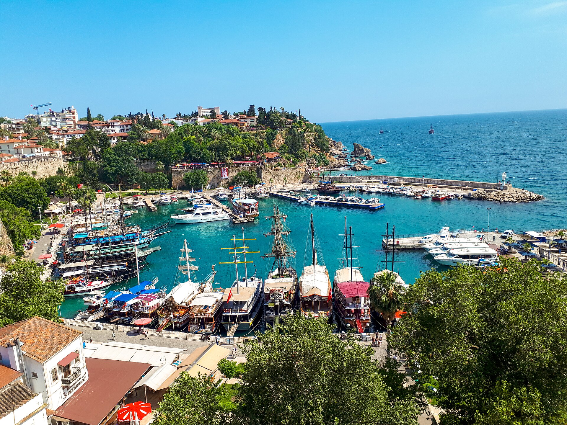 High view over the old Roman harbour of Kaleiçi, Antalya: turquoise water enclosed by stone quays, excursion boats and yachts at the pier, Ottoman houses with red tile roofs climbing the hill behind, the open Mediterranean beyond the breakwater.