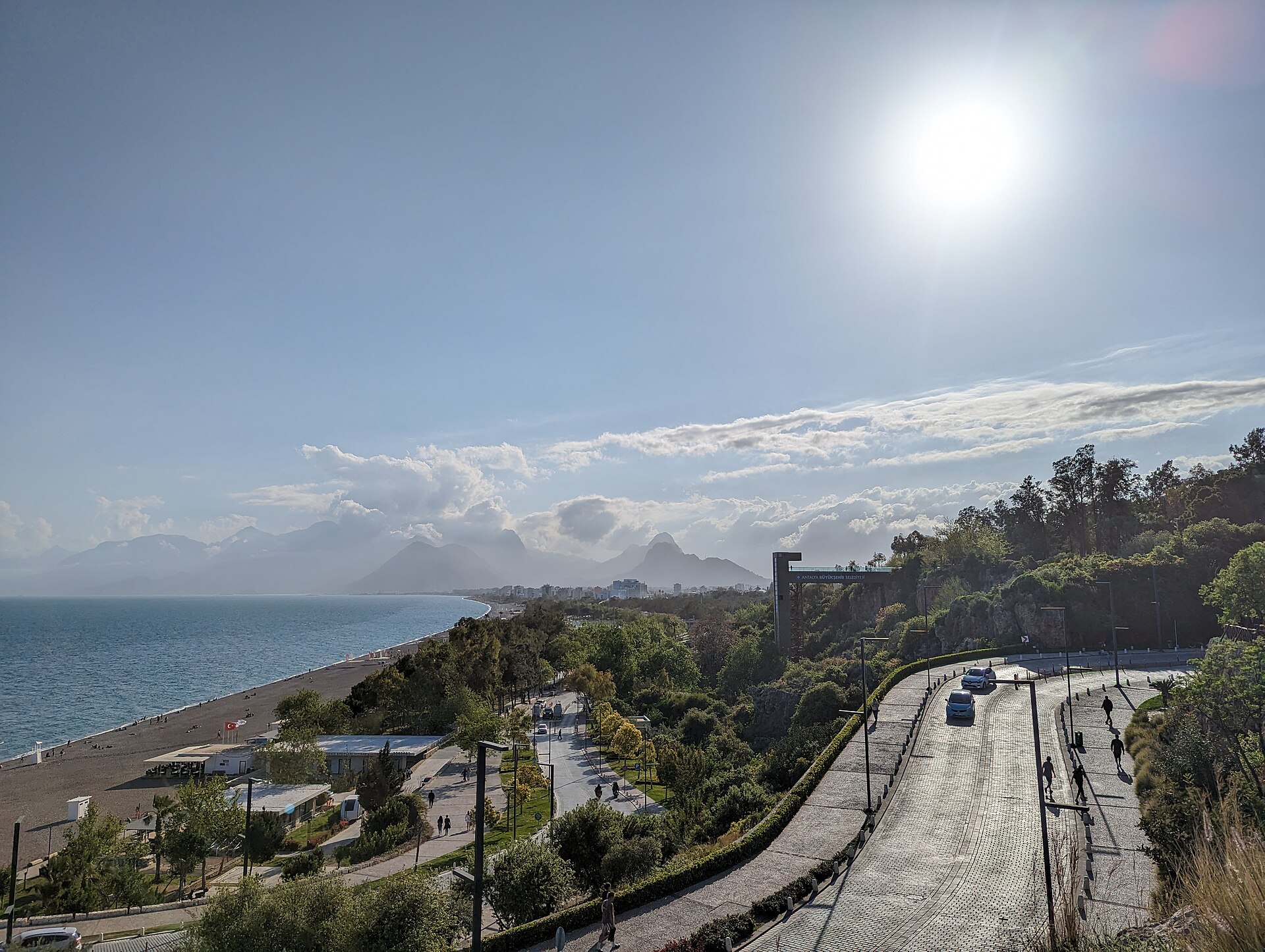 Konyaaltı Beach from the elevated coast road: the grey shingle strand and calm blue-green Mediterranean below, the layered blue ridges of the Beydağları (Taurus) mountains closing the bay.
