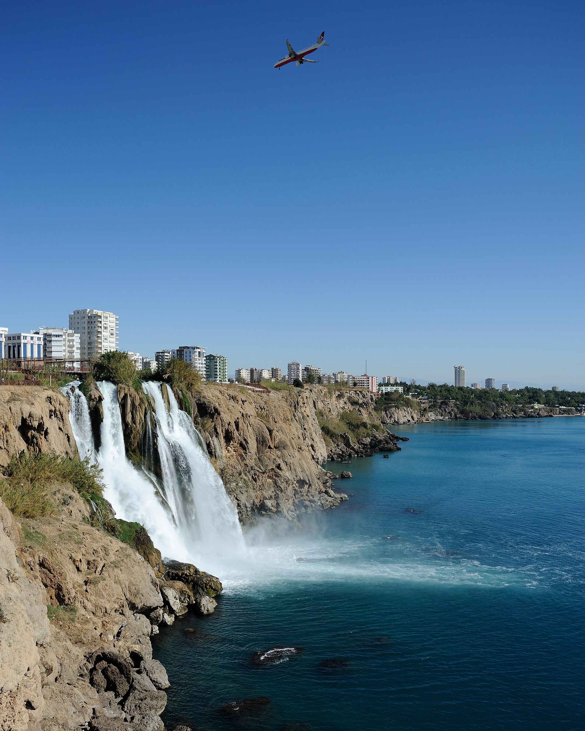 The Lower Düden Falls seen from below the cliffs: white water plunging off the falez into the Mediterranean, spray and rock, open sea beyond.