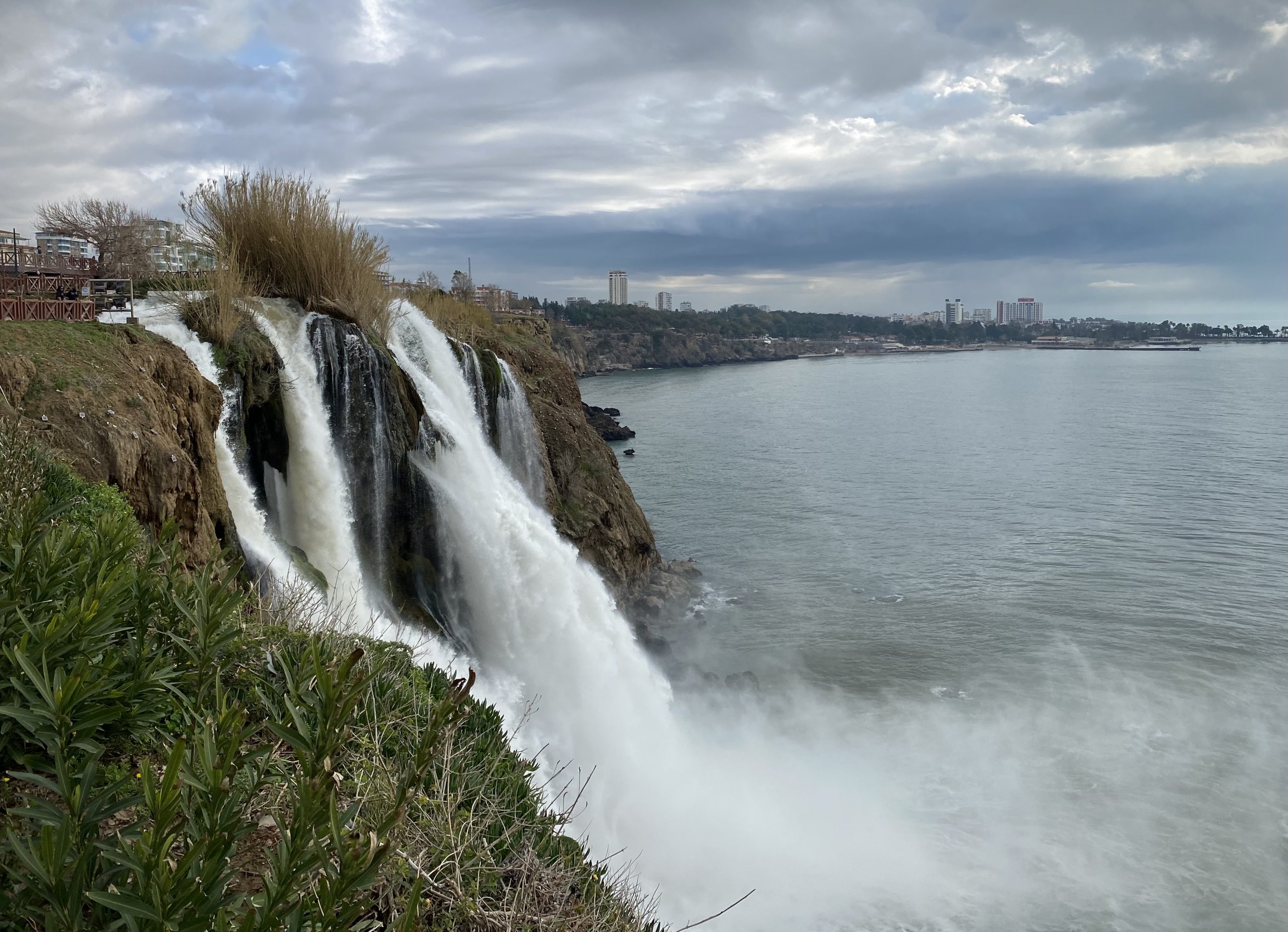 The Lower Düden Falls pouring in white curtains off the falez cliffs directly into the choppy Mediterranean, spray rising, oleander in the foreground, the Antalya skyline far along the coast.