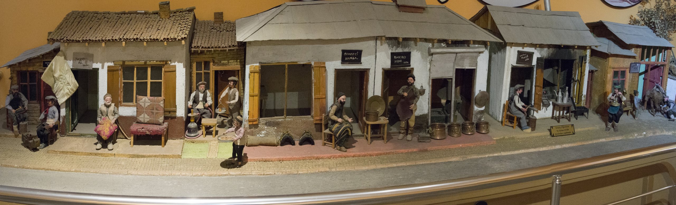 Panorama of the Antalya Archaeological Museum's diorama of old town life: miniature shops with coppersmiths, tailors and tea drinkers under warm golden light.
