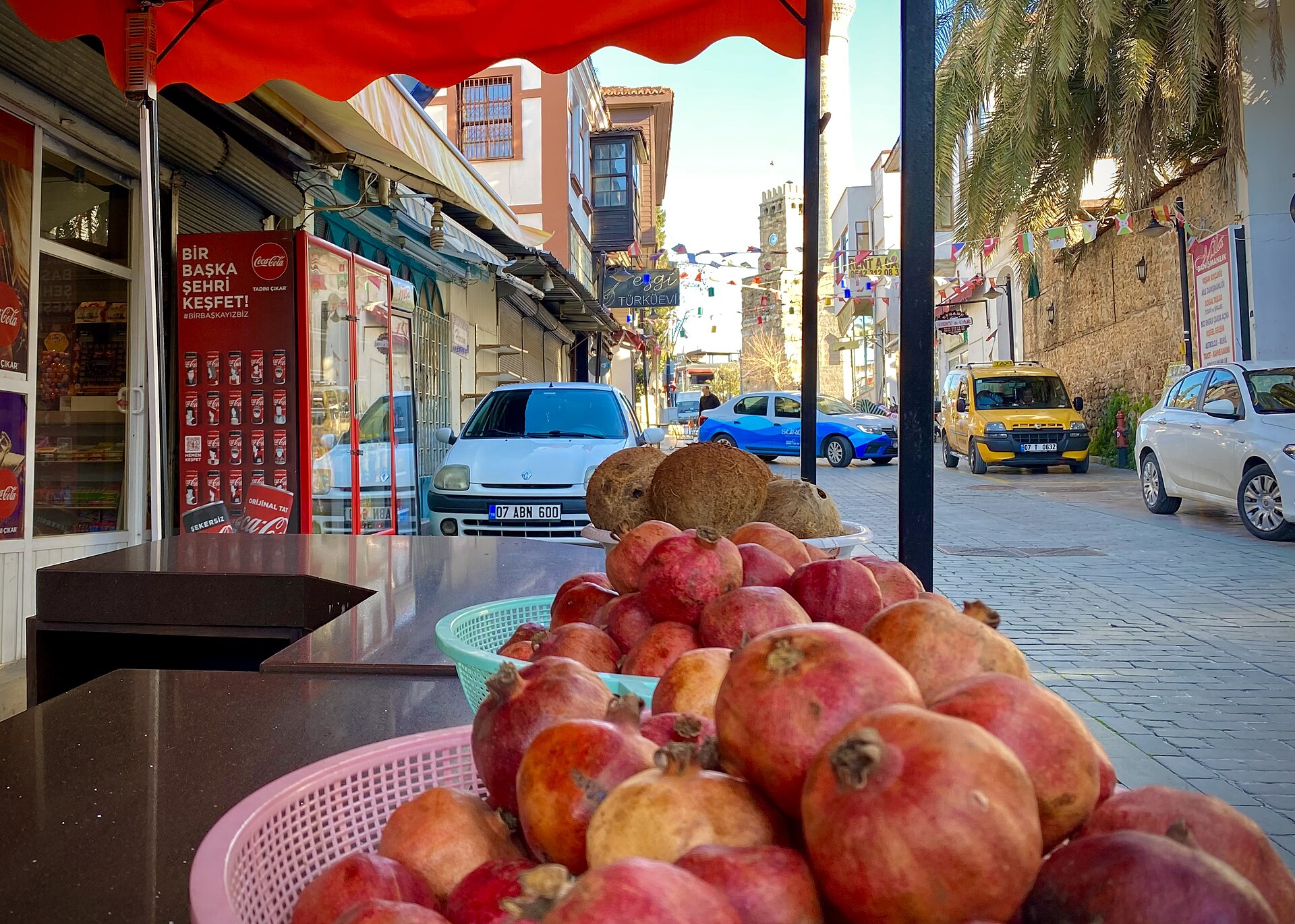 A market display in Kaleiçi: baskets of red and yellow pomegranates on a dark stone counter under a red awning, the old stone clock tower and flags at the end of the cobbled lane.