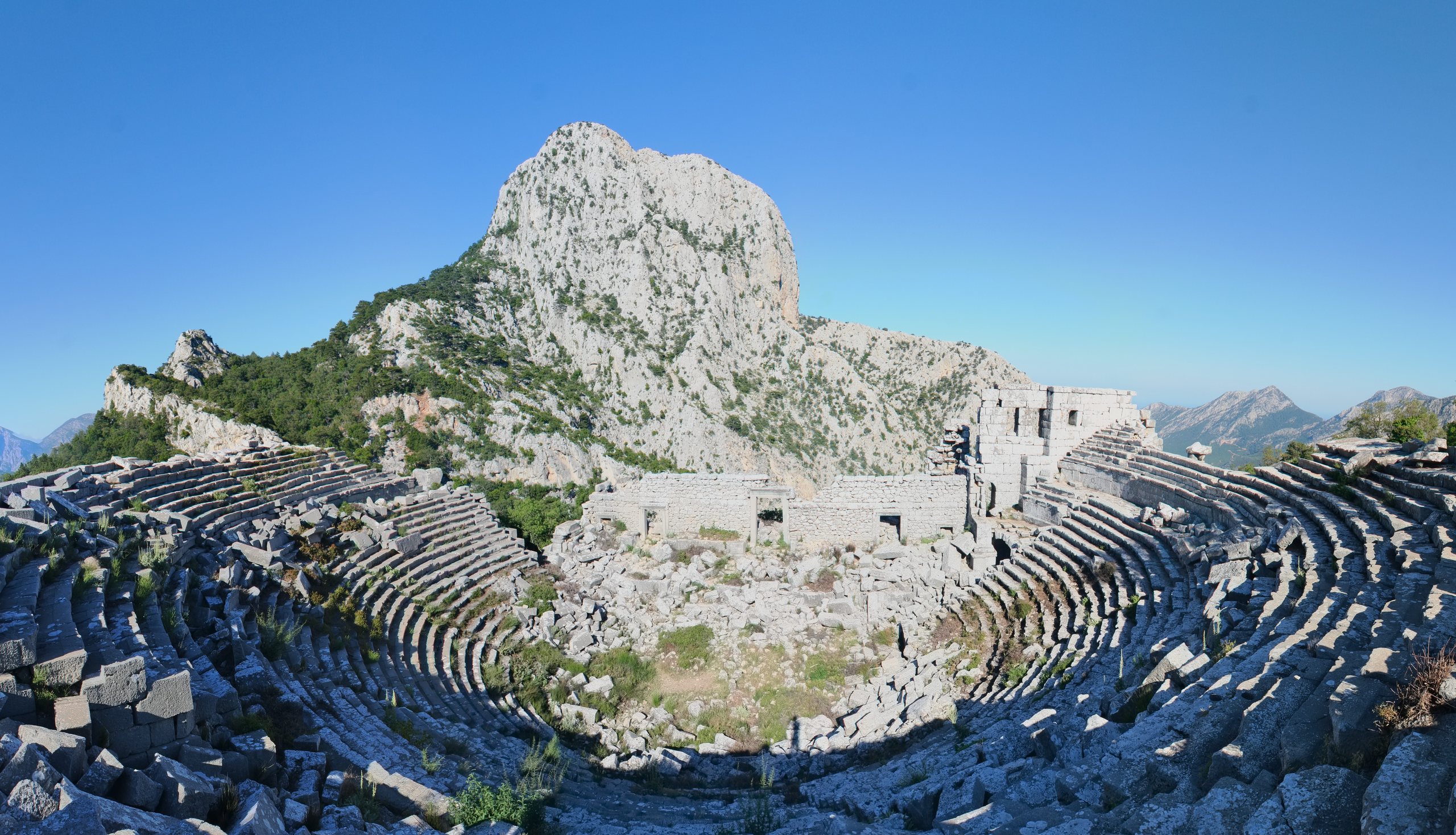 The theatre of Termessos high in the mountains: ruined stone seating, a partly standing stage wall, and the great limestone cliff of Güllük Dağı rising behind against a deep blue sky.