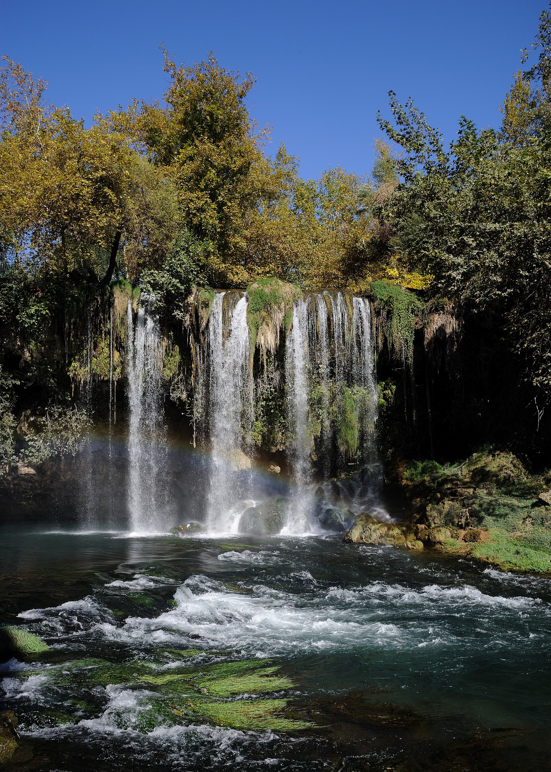 The Upper Düden Falls at Düdenbaşı: water cascading over a mossy travertine ledge into a churning pool, autumn trees and vines hanging over the cliff, a rainbow in the spray.