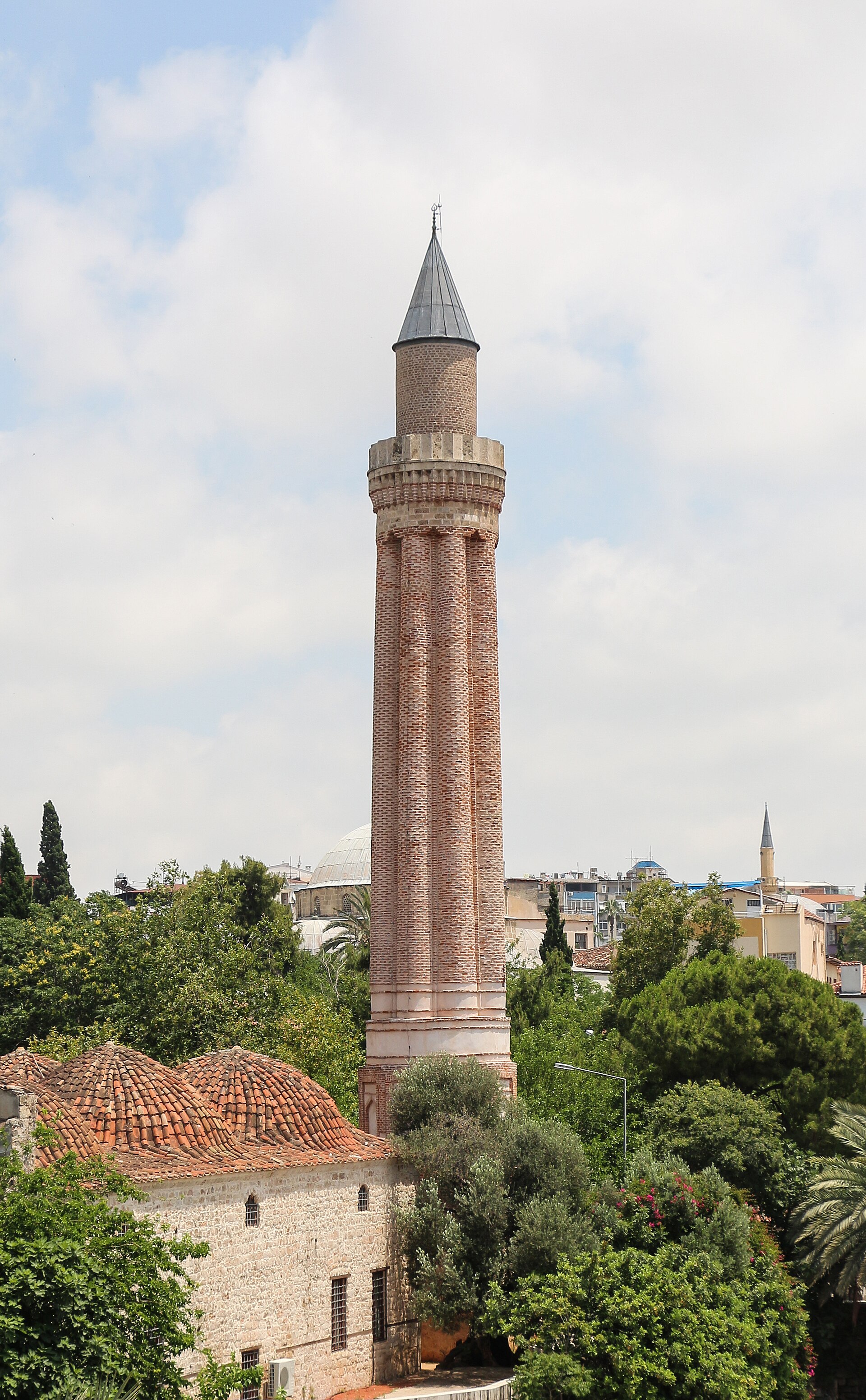The Yivli Minare, Antalya's fluted Seljuk minaret: a tall grooved brick shaft with stone balconies and a dark conical cap, domed mosque buildings and cypresses at its base, modern city behind.
